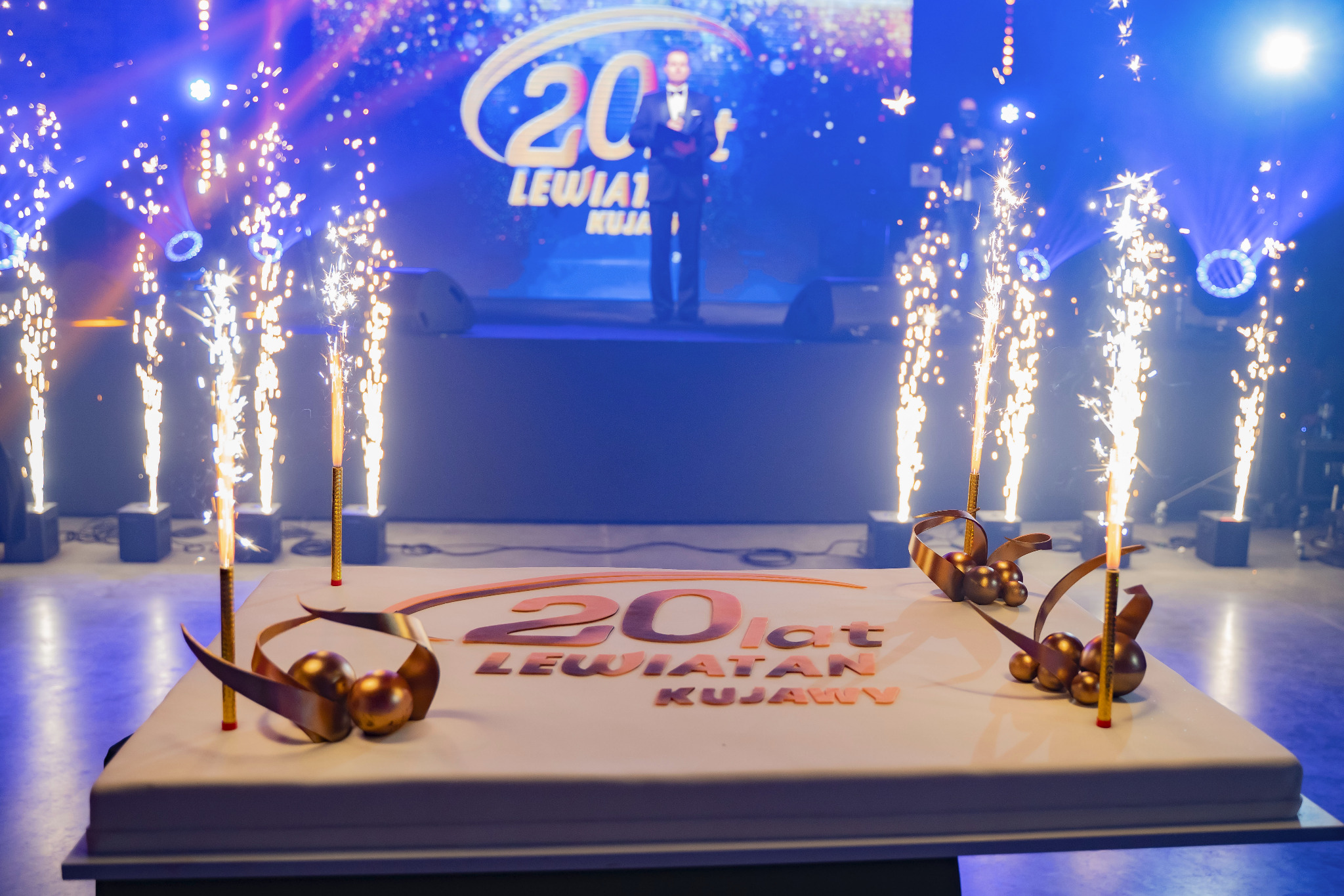 Udekorowany tort z napisem '20 lat Lewiatan Kujawy', fontanny iskier, scena z konferansjerem w tle, oświetlenie sceniczne. Dekoracja urodzinowa na evencie firmowym.