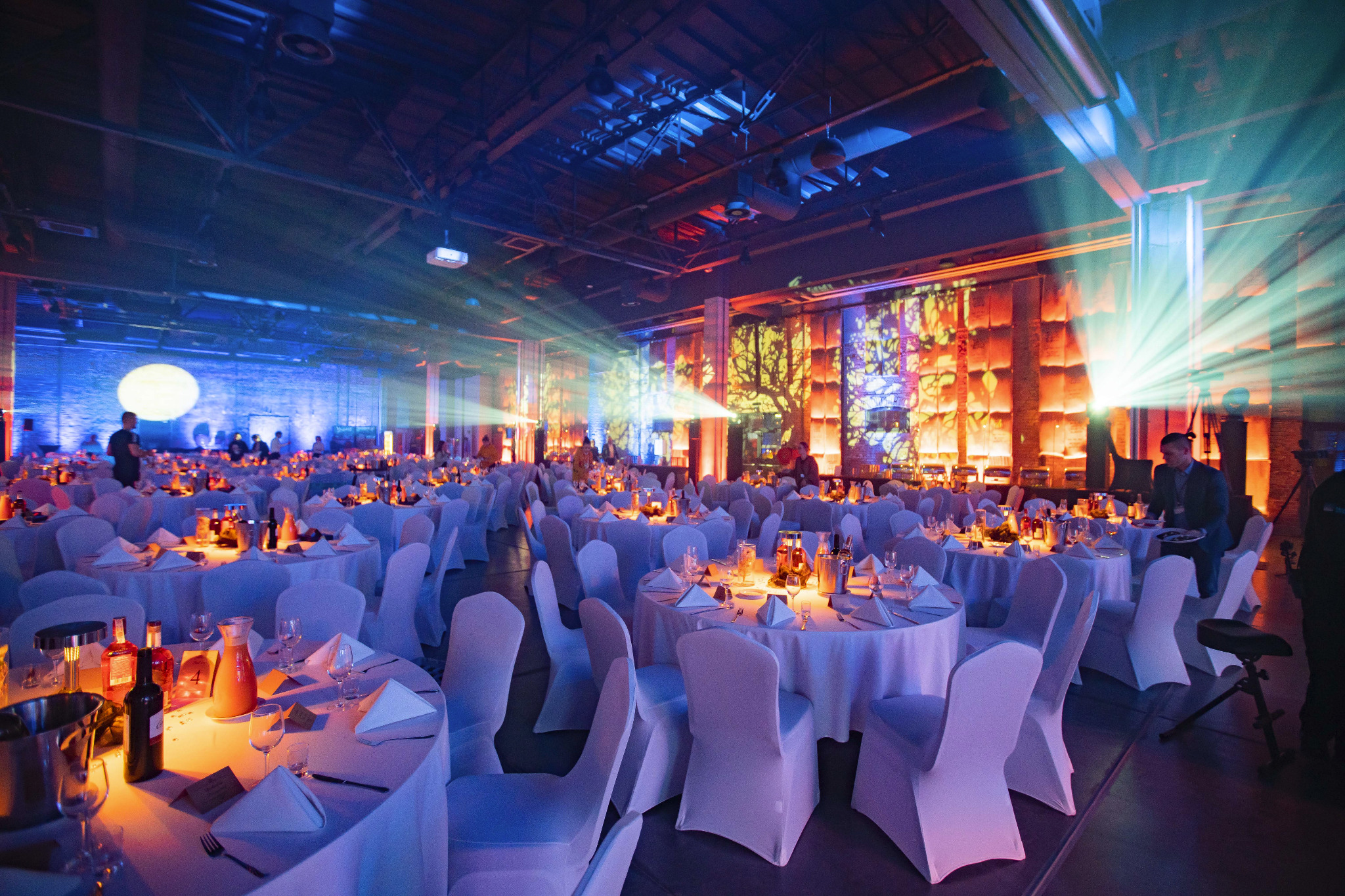 Uroczysta sala bankietowa z okrągłymi stołami w białych obrusach i krzesłach, oświetlona reflektorami i dekoracjami świetlnymi, gotowa na przyjęcie gości.
