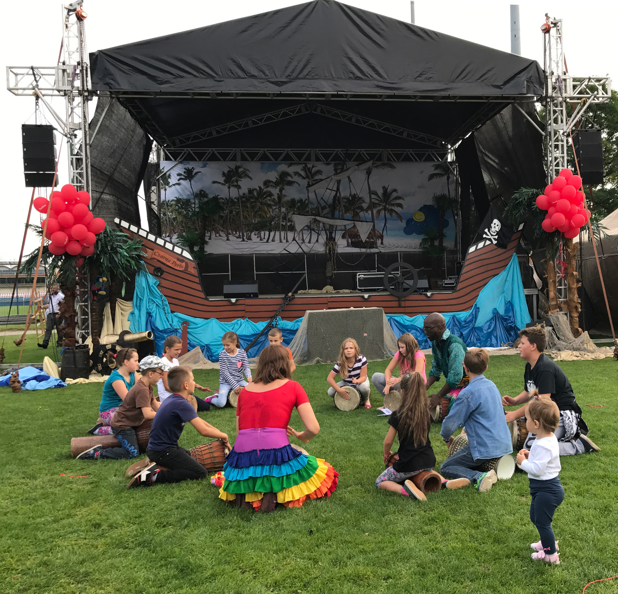 Dzieci i dorośli siedzą na trawie w kręgu, grając na bębnach djembe. W tle scena udekorowana motywem pirackim z namalowaną scenerią tropikalnej wyspy i elementami statku Czarna Perła.