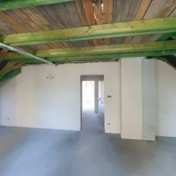 Wnętrze w trakcie prac wykończeniowych: świeżo otynkowane ściany, surowy drewniany strop z zielonymi belkami, widoczne instalacje elektryczne i otwory drzwiowe.