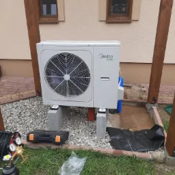 Zewnętrzna jednostka pompy ciepła Midea R32, zamontowana na betonowych bloczkach na żwirowym podłożu przy domu, z widocznymi elementami instalacji i narzędziami w tle.