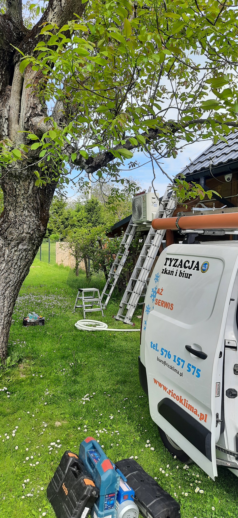 Montaż jednostki zewnętrznej klimatyzacji na elewacji domu, widok z poziomu trawnika z narzędziami i samochodem serwisowym firmy Ricoklima.pl
