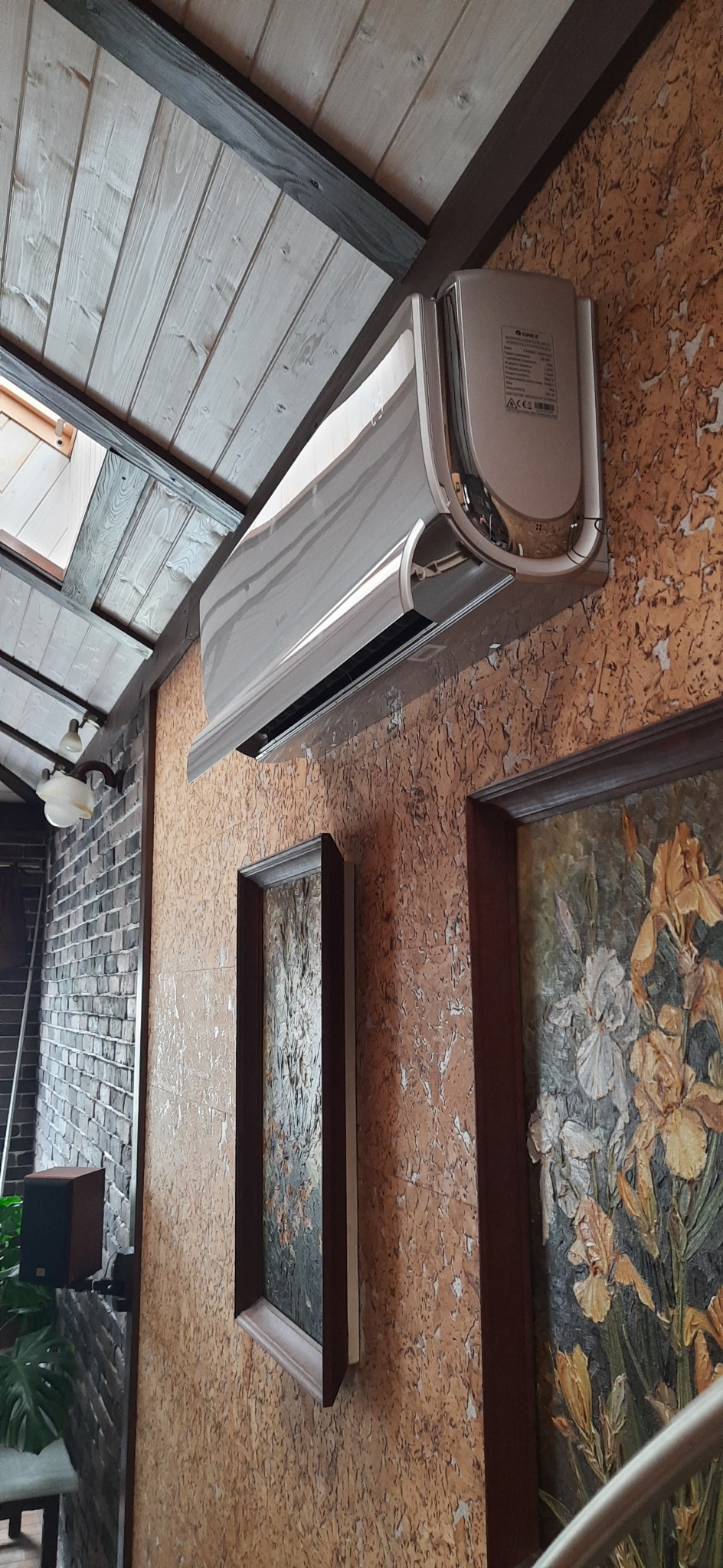 Srebrny klimatyzator zamontowany na ścianie o fakturze kory drzewa, wiszący nad dwoma obrazami w ciemnych ramach, z widocznym fragmentem sufitu z drewnianych desek i lampą w tle.