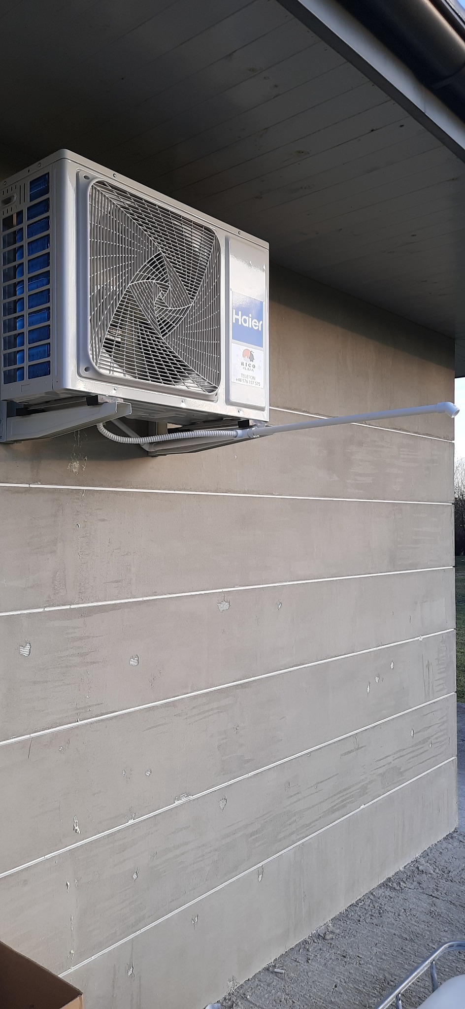 Zamontowana jednostka zewnętrzna klimatyzacji Haier z odprowadzeniem skroplin przy ścianie budynku wykończonej poziomymi panelami.