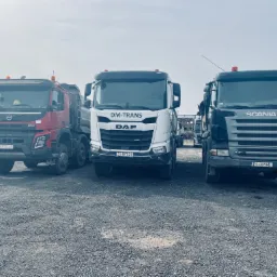 Trzy ciężarówki: czerwony Volvo, biały DAF z logo DM-Trans i szary Scania, stoją na placu budowy. W tle widoczne elementy konstrukcyjne i budynki.