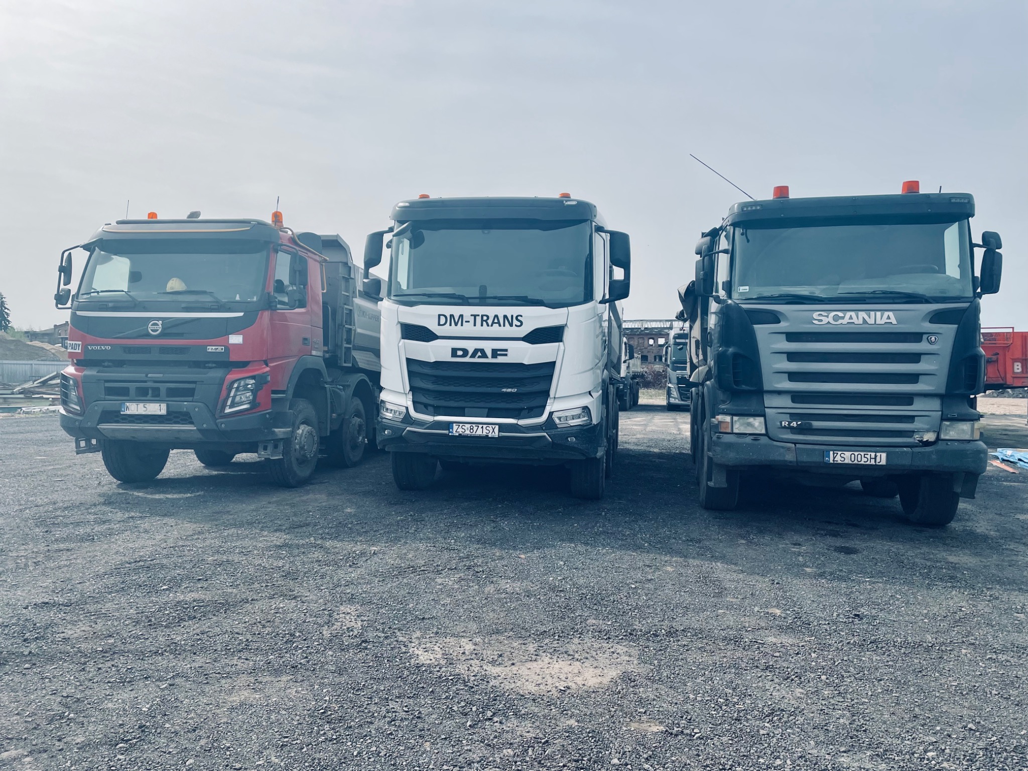 Trzy ciężarówki: czerwony Volvo, biały DAF z logo DM-Trans i szary Scania, stoją na placu budowy. W tle widoczne elementy konstrukcyjne i budynki.