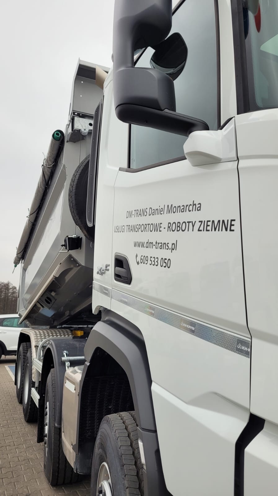 Ciężarówka z wywrotką, z widocznym logo firmy DM-TRANS Daniel Monarcha, oferującej usługi transportowe i roboty ziemne. Widoczny numer telefonu i adres strony internetowej.
