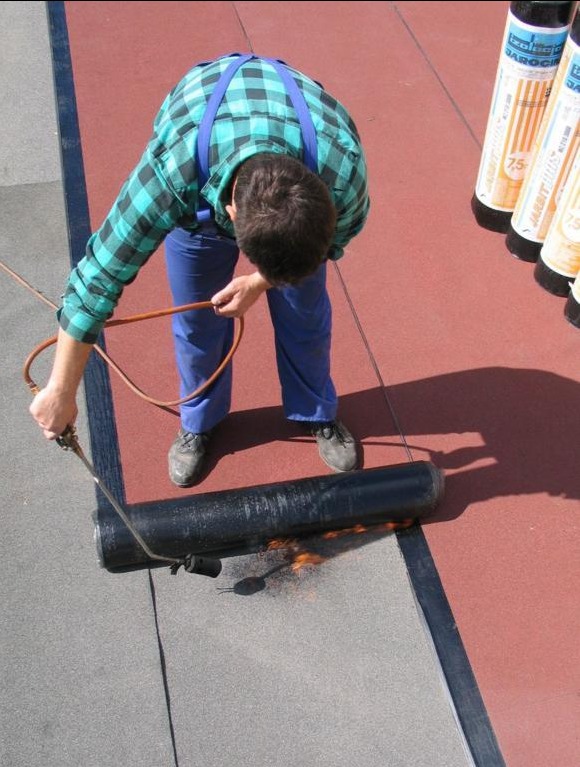 Dekarz w niebieskiej kraciastej koszuli i spodniach, z góry, zgrzewa papę na dachu za pomocą palnika, widoczne rolki papy w tle.