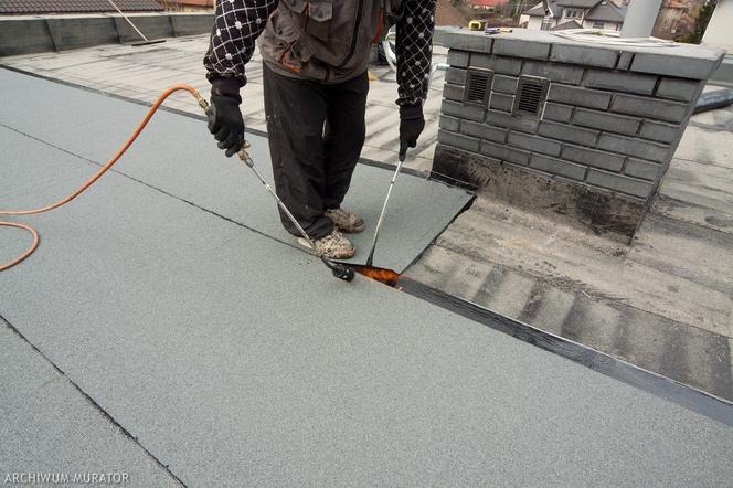 Pracownik w kombinezonie spawa papę termozgrzewalną na dachu, widoczny płomień palnika i fragment komina z cegły