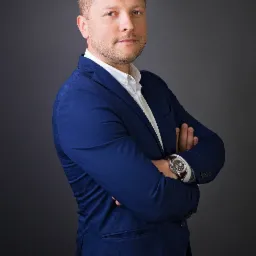 Portret mężczyzny w granatowym garniturze i białej koszuli, z zegarkiem na skórzanym pasku, stojącego na tle grafitowego tła z założonymi rękami.