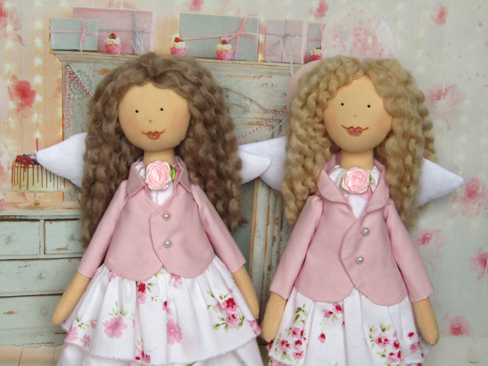 Dwie ręcznie robione lalki szmaciane anioły w pastelowych różowych marynarkach i kwiecistych sukienkach, stojące na tle dekoracji urodzinowych.