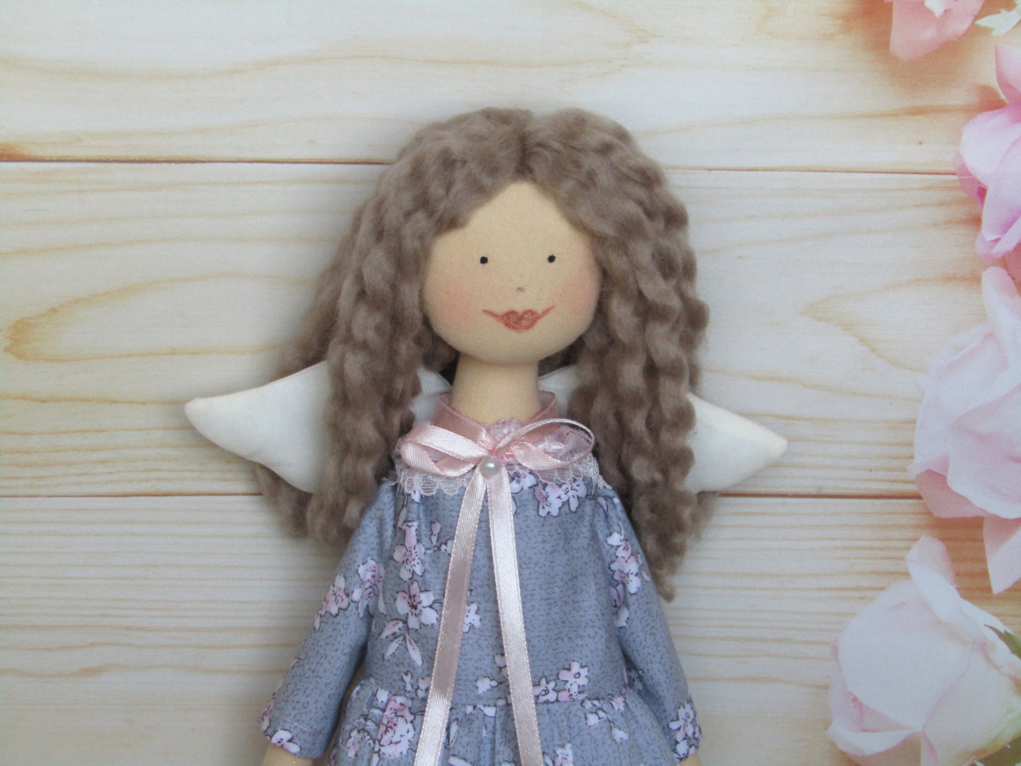 Ręcznie robiona lalka szmaciana z kręconymi włosami, w sukience w kwiaty i z białymi skrzydłami, ozdobiona różową wstążką, na drewnianym tle z kwiatami.