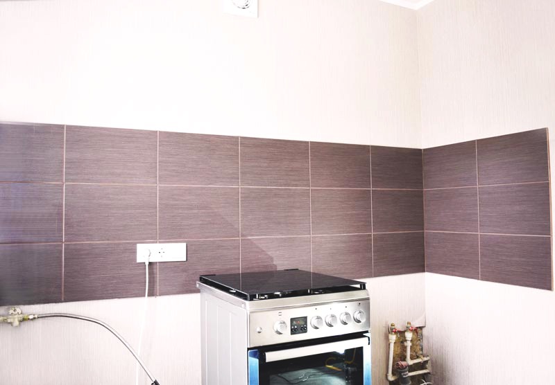 Kuchnia w trakcie remontu: ułożone brązowe płytki imitujące drewno nad kuchenką gazową, widoczne gniazdko elektryczne i rury doprowadzające gaz.