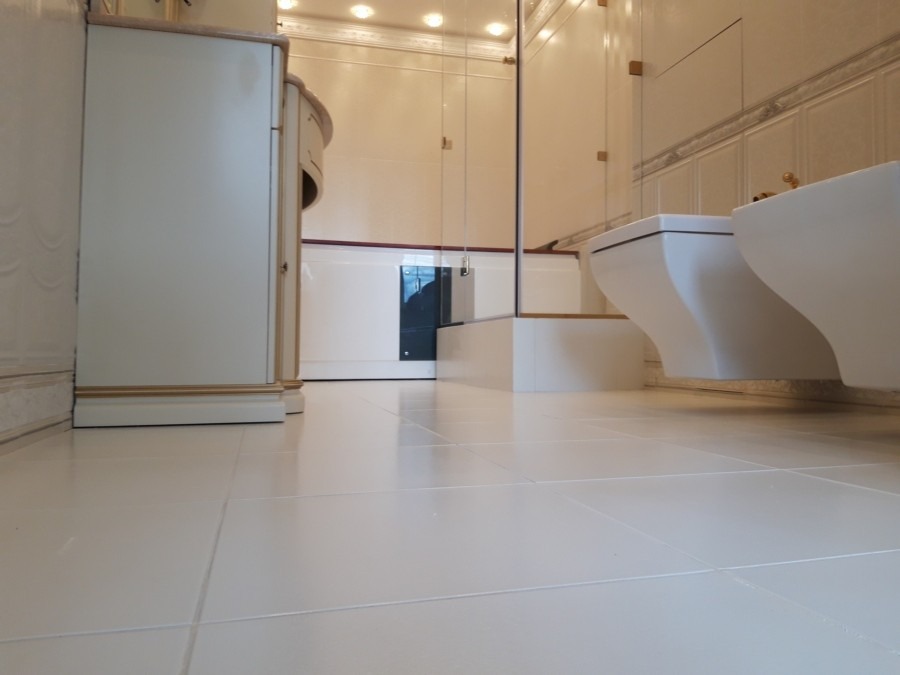 Jasna, luksusowa łazienka z białymi płytkami podłogowymi, wiszącą toaletą i bidetem, oraz szafką z beżowymi akcentami, widok z niskiej perspektywy podkreślający połysk płytek.