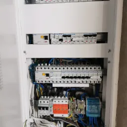 Otwarta skrzynka bezpiecznikowa z zainstalowanymi wyłącznikami EATON, zabezpieczeniami przepięciowymi PRO-TEC i modułami SIMET, widoczne różnokolorowe przewody elektryczne.