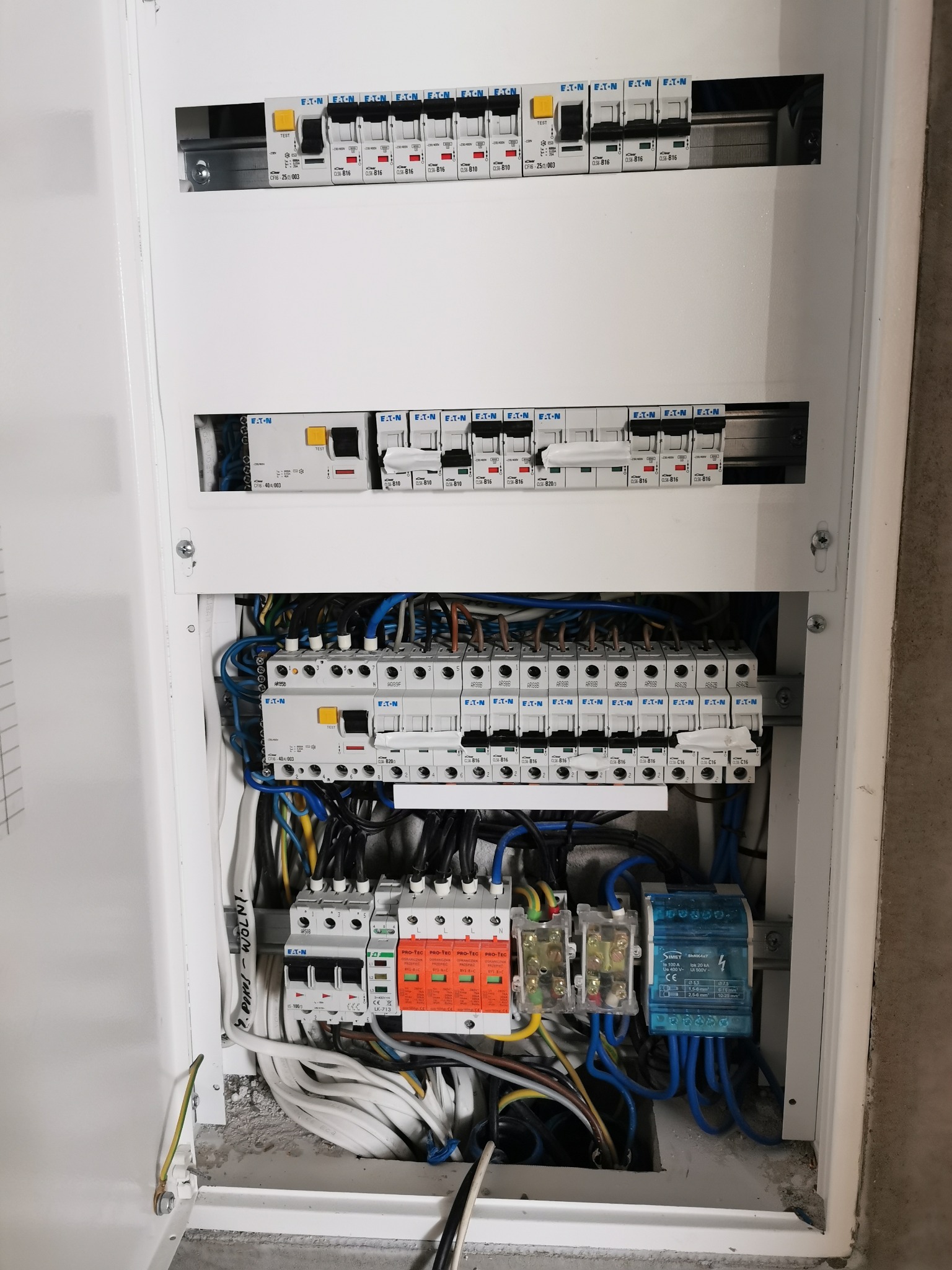 Otwarta skrzynka bezpiecznikowa z zainstalowanymi wyłącznikami EATON, zabezpieczeniami przepięciowymi PRO-TEC i modułami SIMET, widoczne różnokolorowe przewody elektryczne.