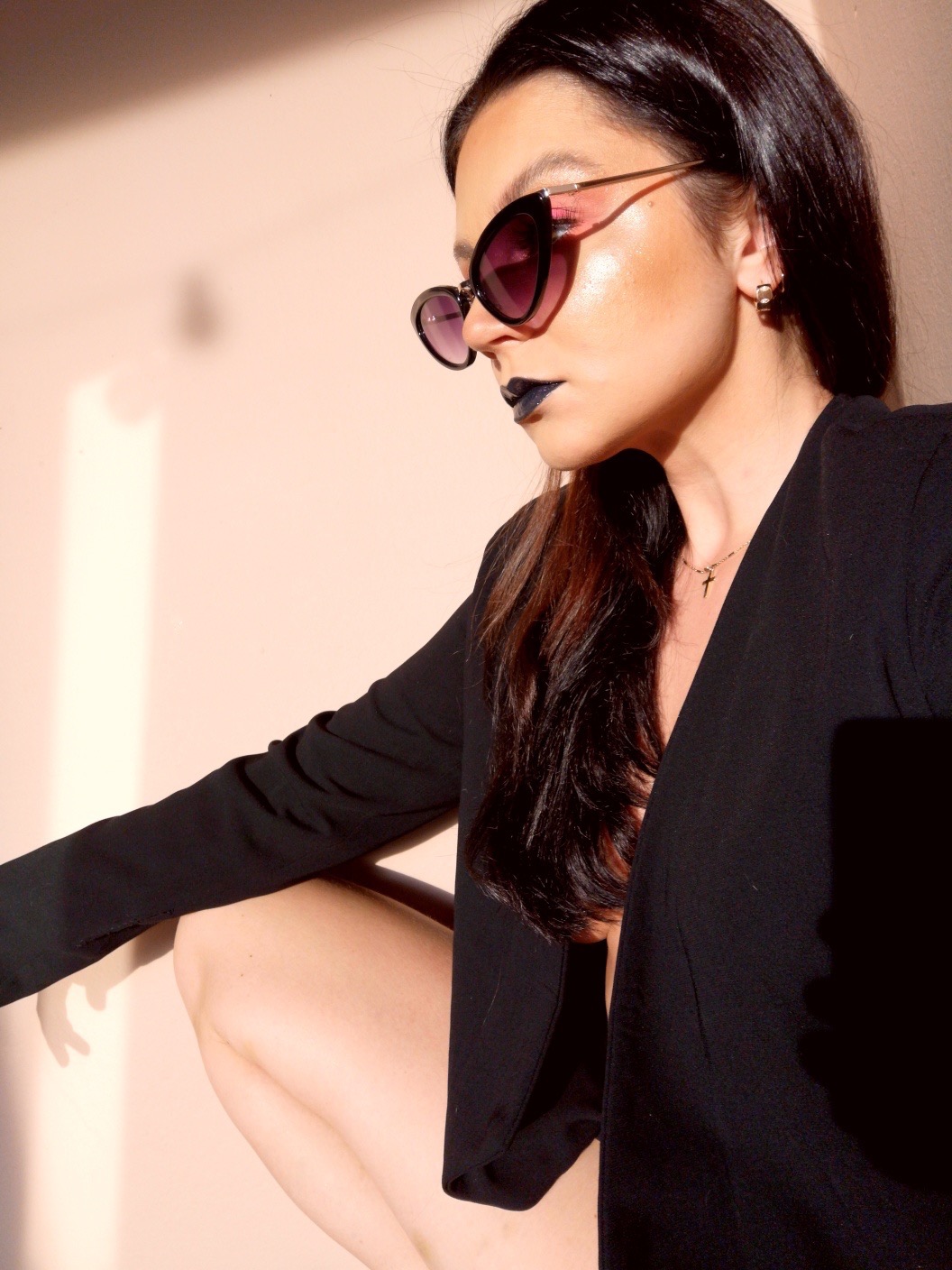 Profil kobiety w okularach przeciwsłonecznych, z ciemną szminką i błyszczącym makijażem, ubrana w czarny żakiet.
