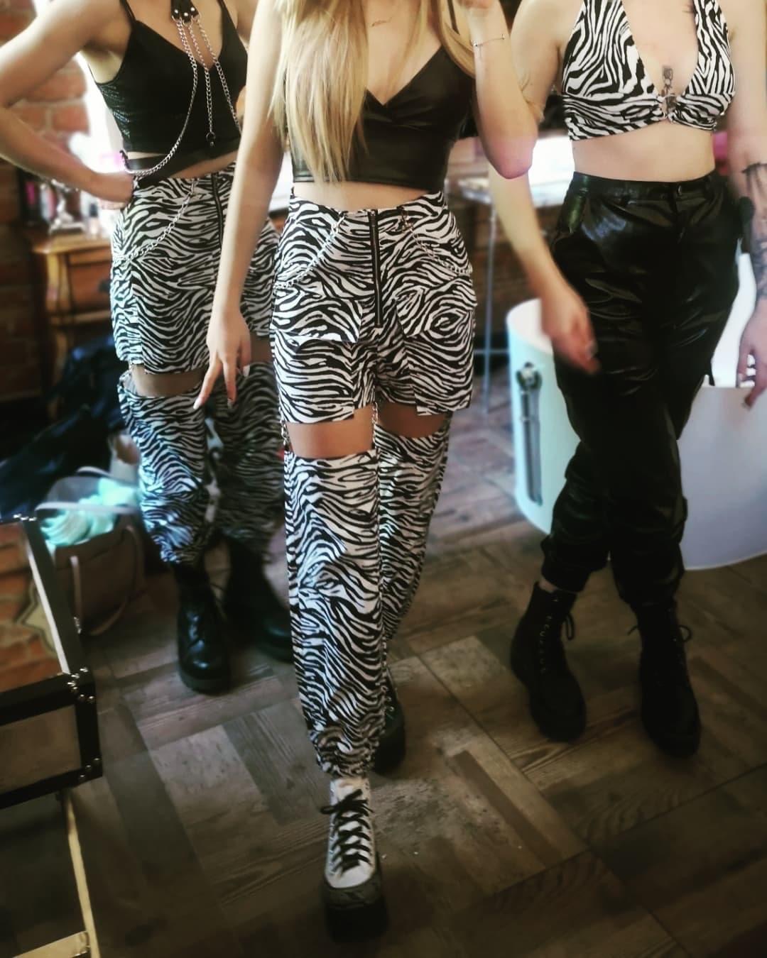 Trzy kobiety prezentują stylizacje: skórzany top z łańcuszkami, spodnie i szorty w zebrę z łańcuszkami, czarne skórzane spodnie i top w zebrę, czarne buty na platformie.