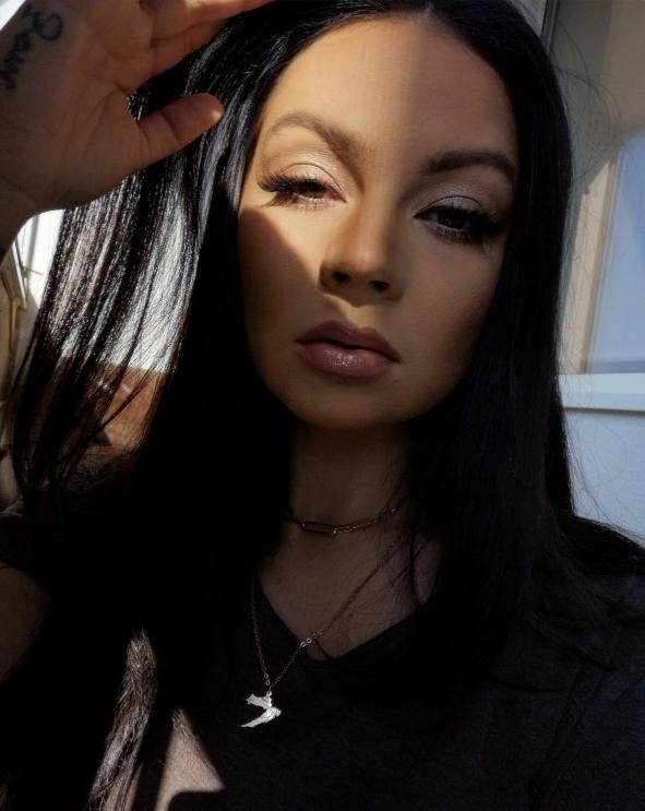 Portret kobiety z mocnym makijażem oczu i ustami w odcieniu nude, oświetlonej promieniami słońca, z widocznym tatuażem na dłoni i delikatnym naszyjnikiem z zawieszką w kształcie ptaka.