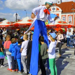 Mężczyzna w kostiumie na szczudłach, w białym cylindrze i z czerwoną muszką, występuje dla grupy dzieci na tle miejskiego placu z parasolami piwnymi Tyskie.