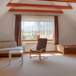 Jasny pokój na poddaszu z drewnianymi belkami, beżową kanapą narożną, drewnianym krzesłem, łóżkiem i oknem z zasłonami.