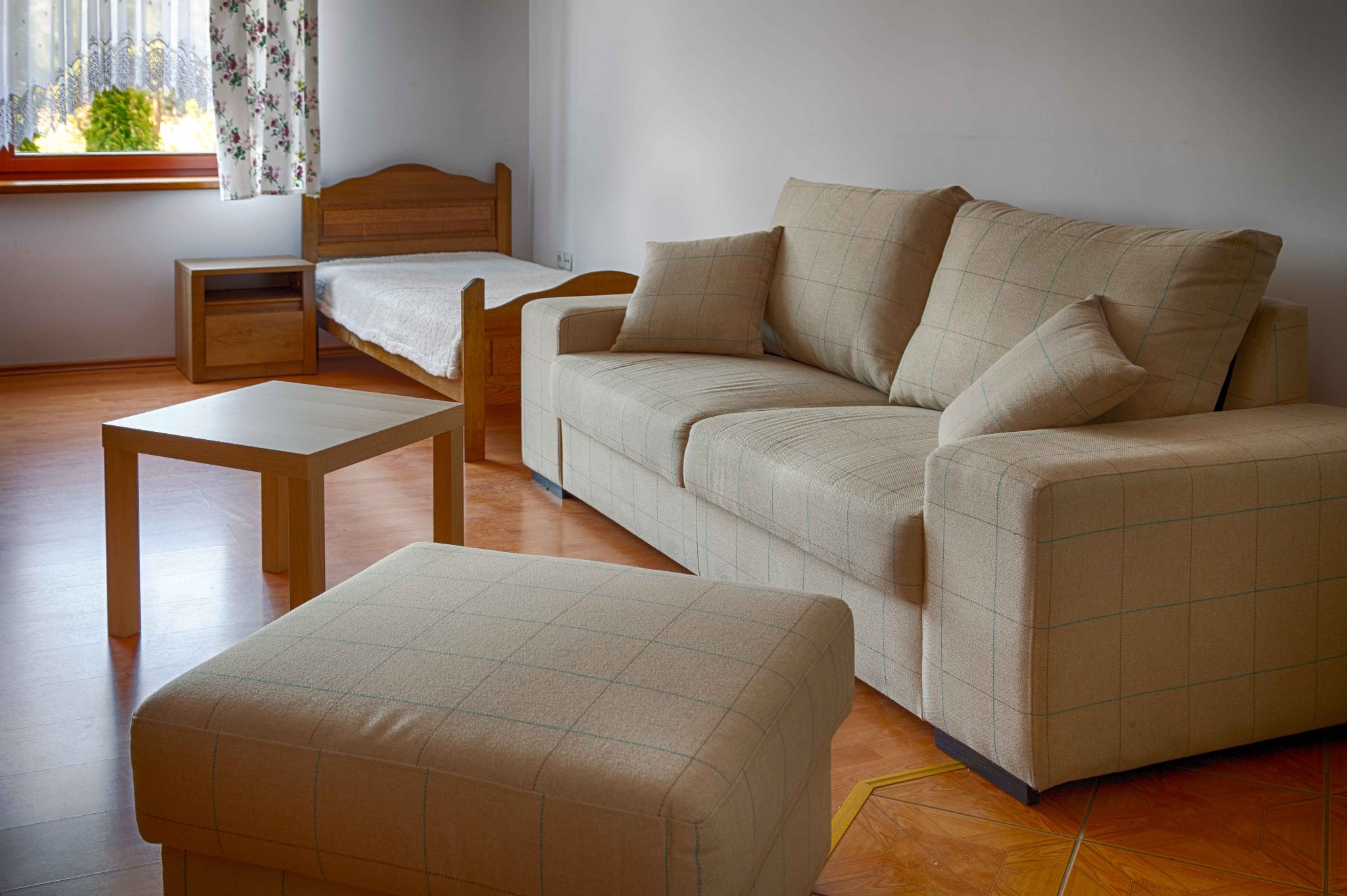 Pokój w jasnych barwach z drewnianym łóżkiem, stolikiem i beżową sofą w kratę z podnóżkiem, oświetlony przez okno z kwiecistą firanką.