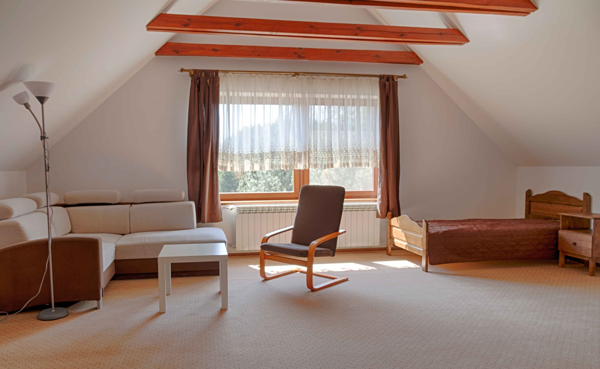 Jasny pokój na poddaszu z drewnianymi belkami, beżową kanapą narożną, drewnianym krzesłem, łóżkiem i oknem z zasłonami.