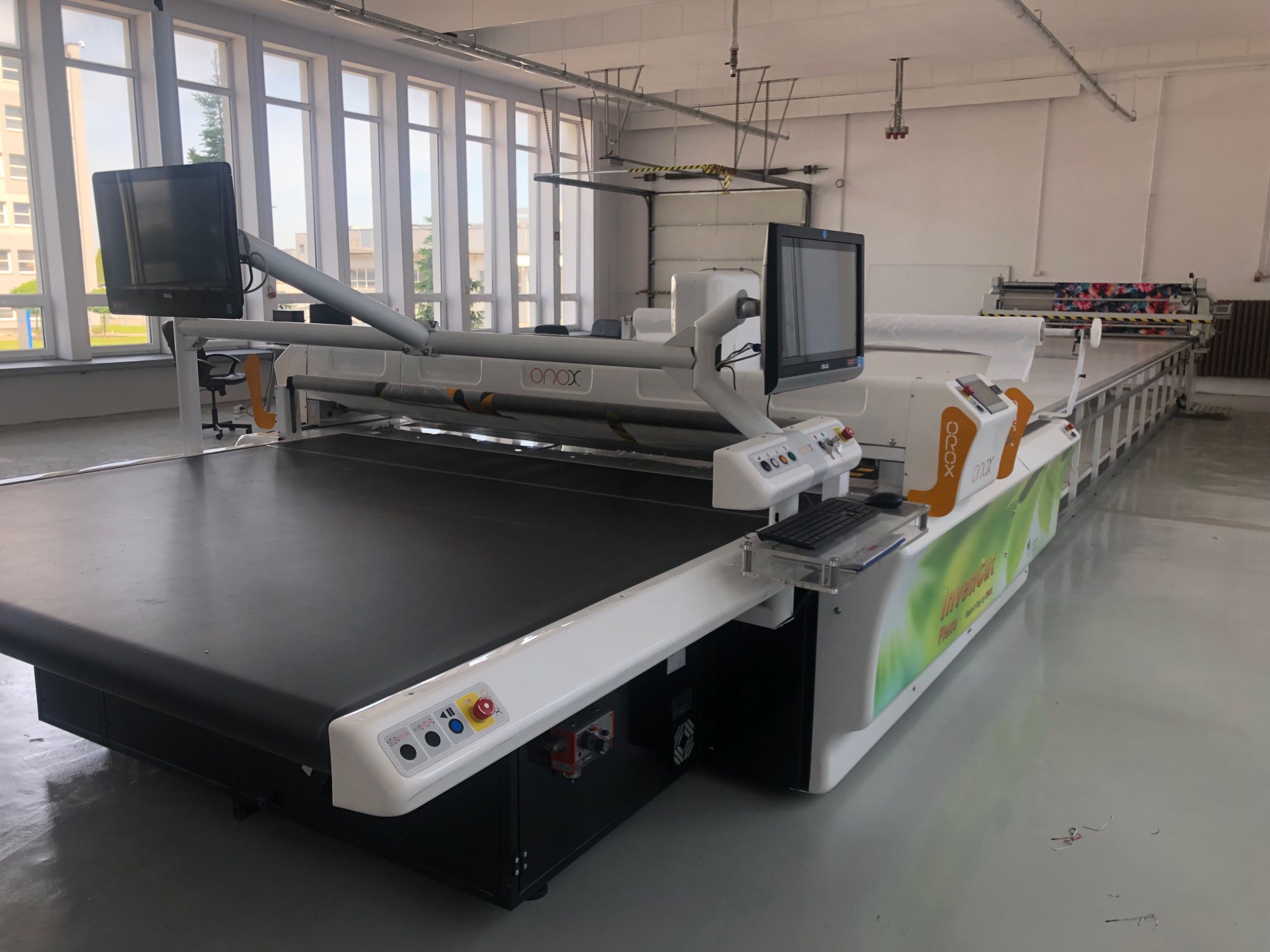 Długa, nowoczesna maszyna krojcza z ekranami i klawiaturą w jasnej hali produkcyjnej. Widoczny system podawania materiału i elementy sterujące.