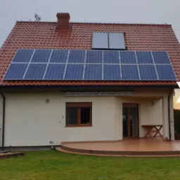 Dom jednorodzinny z czerwonym dachem i panelami słonecznymi na dachu, widoczne również kolektory słoneczne.