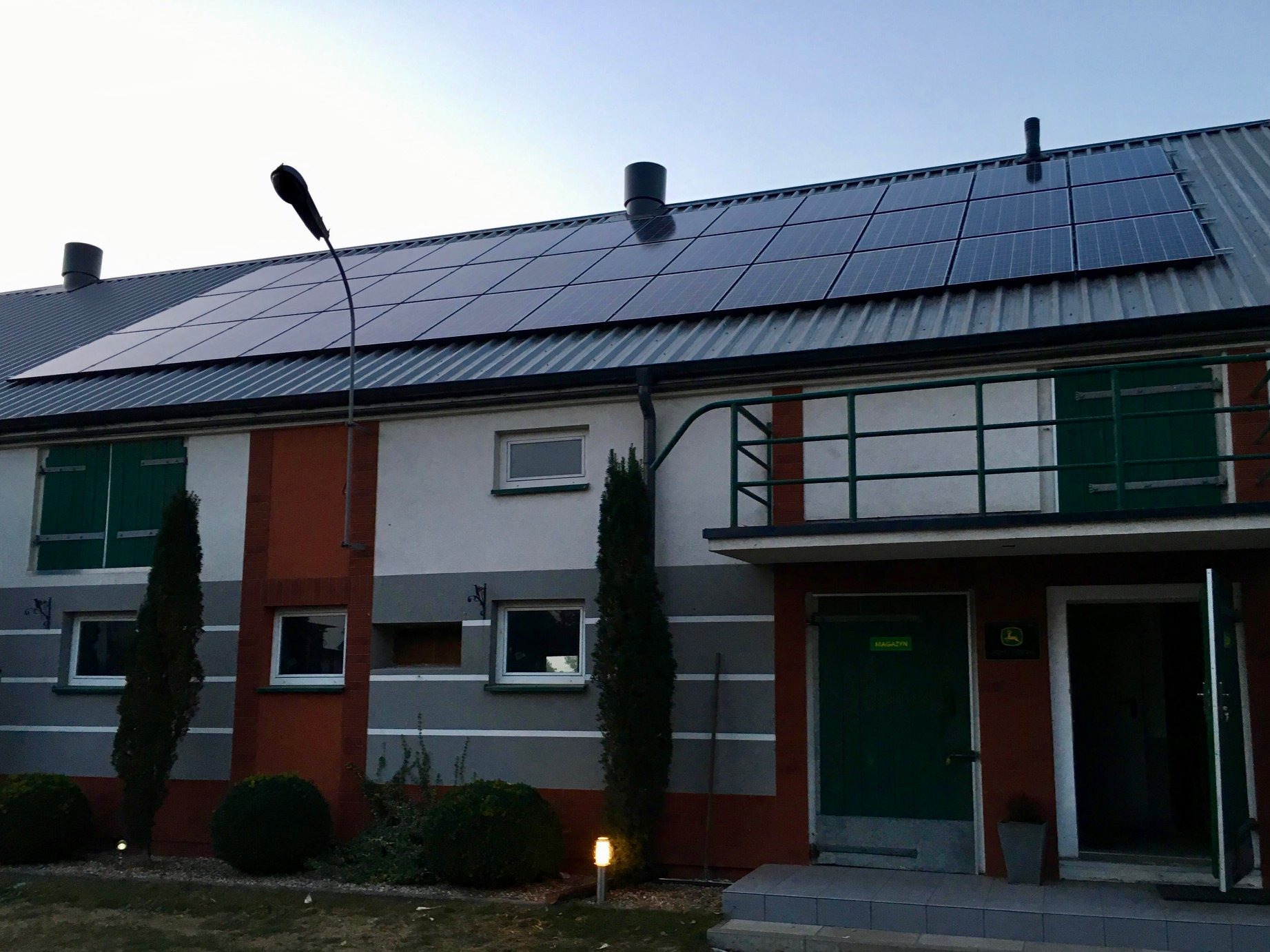 Panel słoneczny na dachu budynku gospodarczego z zielonymi drzwiami i elementami dekoracyjnymi, oświetlony lampą uliczną.