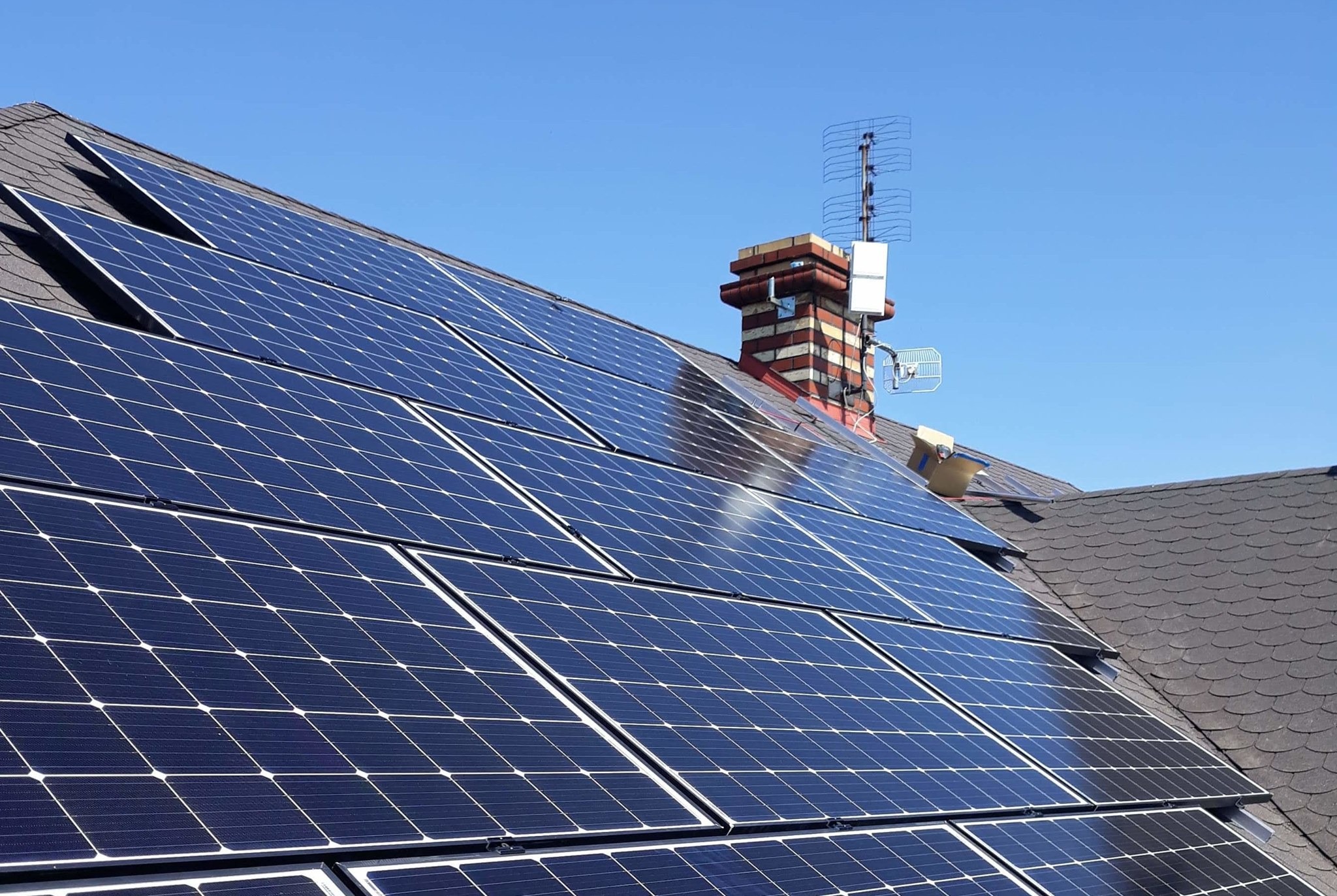 Panele słoneczne zamontowane na dachu domu z ceglanym kominem i anteną telewizyjną w tle, widoczny karton na dachu.