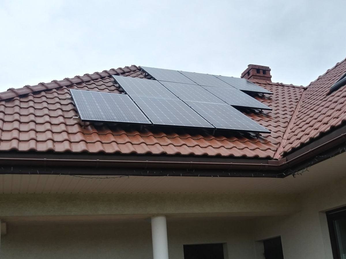 Panele fotowoltaiczne zamontowane na dachu z czerwonej dachówki, widoczny komin i fragment elewacji budynku.