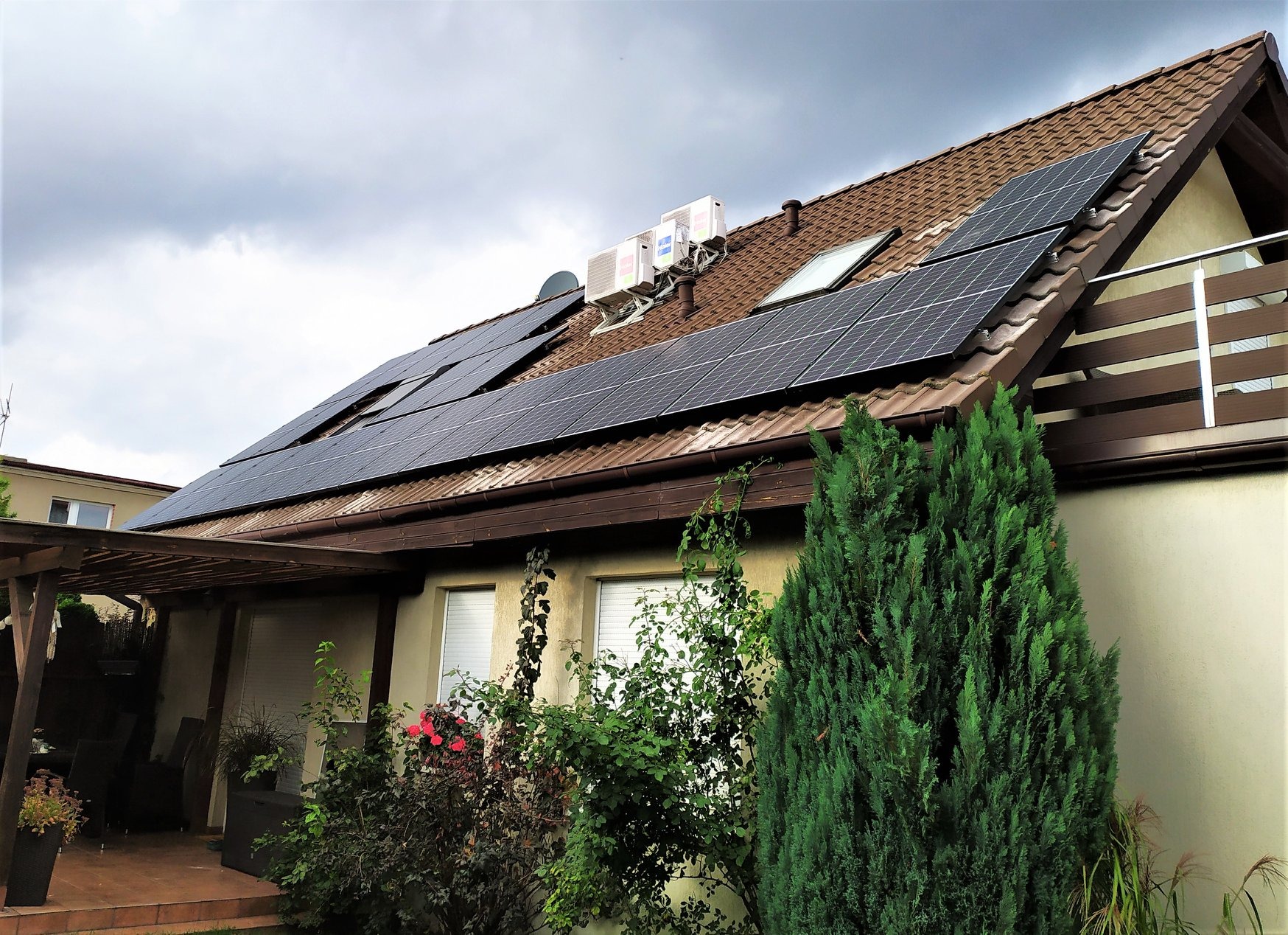 Panele słoneczne na brązowym dachu domu jednorodzinnego, widoczne klimatyzatory i okno dachowe, zieleń w ogrodzie.