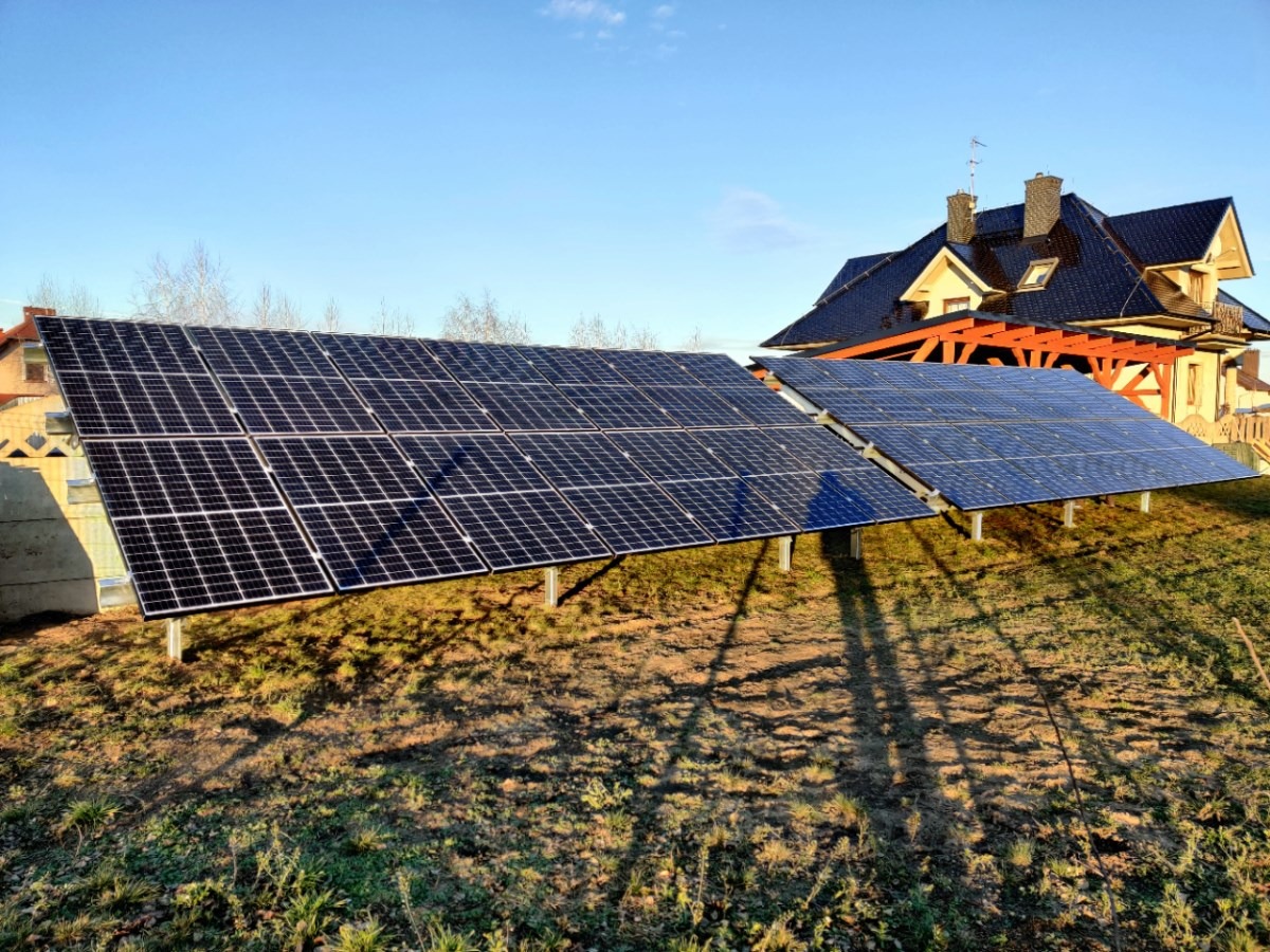 Dwa rzędy paneli słonecznych ustawione na metalowych konstrukcjach na tle domu z ciemnym dachem i błękitnego nieba. Cienie paneli widoczne na trawie.
