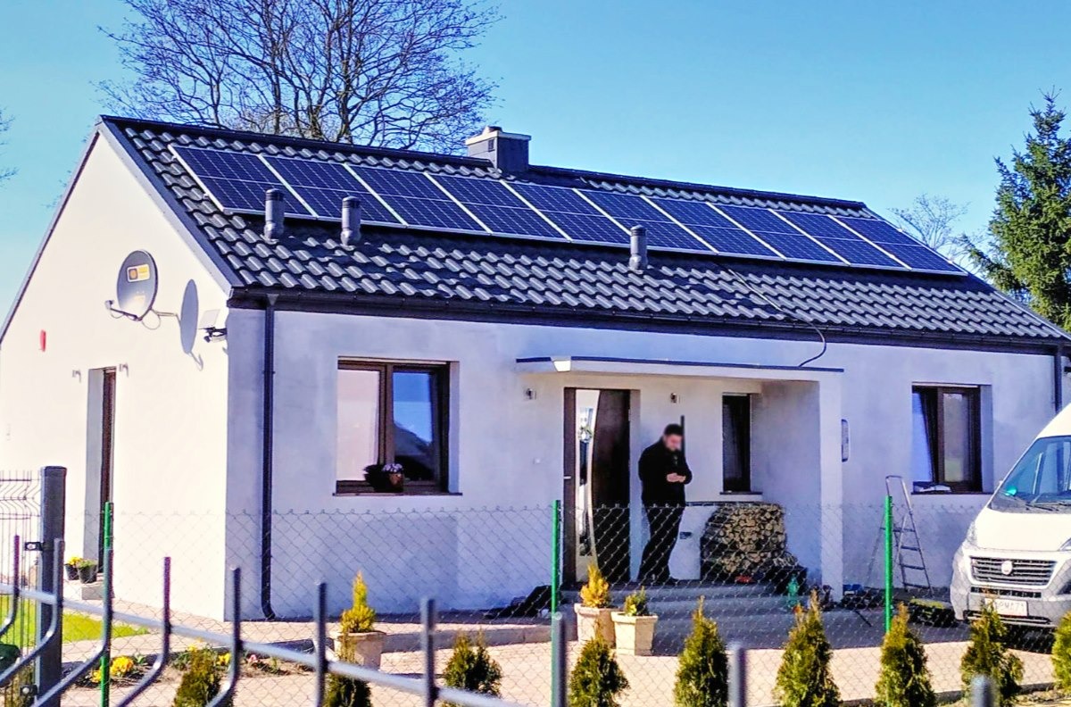 Biały dom jednorodzinny z ciemną dachówką i panelami słonecznymi na dachu; na werandzie stoi mężczyzna, obok zaparkowany biały bus.