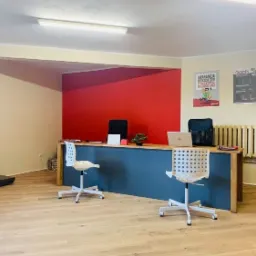 Wnętrze biura firmy transportowej z wagą platformową, biurkiem z laptopem i krzesłami obrotowymi, plakatami informacyjnymi na ścianie i akcentem w postaci czerwonej ściany.