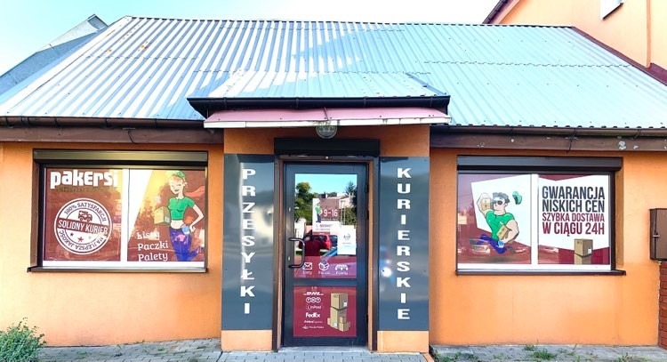 Fasada budynku firmy kurierskiej z napisem 'Przesyłki Kurierskie', logotypami firm spedycyjnych na drzwiach i reklamami w oknach z gwarancją niskich cen i szybkiej dostawy.