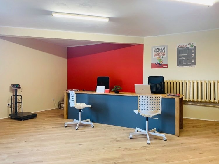 Wnętrze biura firmy transportowej z wagą platformową, biurkiem z laptopem i krzesłami obrotowymi, plakatami informacyjnymi na ścianie i akcentem w postaci czerwonej ściany.