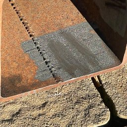 KMW Group Wojciech Rataj - Fragment zardzewiałego, metalowego profilu z widocznym śladem cięcia i obszarem po piaskowaniu, leżącego na piasku. Detal obróbki metalu.