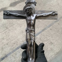 KMW Group Wojciech Rataj - Metalowy krucyfiks trzymany w rękawicy roboczej na tle betonowej posadzki. Widoczne detale faktury metalu i zarys postaci Jezusa.