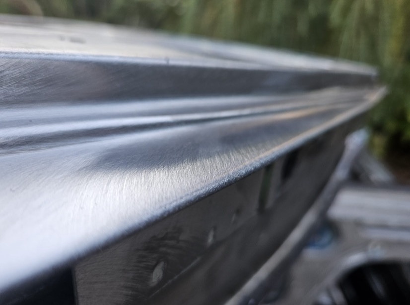 Szlifowana powierzchnia metalowa z widocznymi śladami obróbki, prawdopodobnie element konstrukcyjny lub karoserii pojazdu. Delikatne odbicia światła podkreślają fakturę.