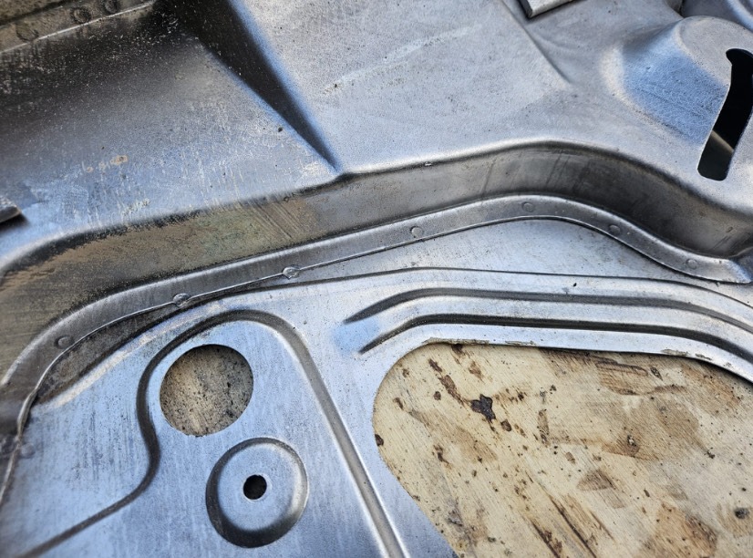 Szczegółowe ujęcie metalowej formy z otworami i przetłoczeniami, leżącej na drewnianej powierzchni. Widoczne ślady obróbki i patyna na metalu.