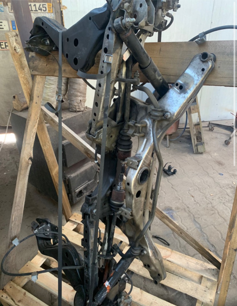 Elementy zawieszenia samochodowego na drewnianym stojaku. Widoczne metalowe ramy, sprężyny i amortyzatory. Częściowo widoczne numery rejestracyjne.