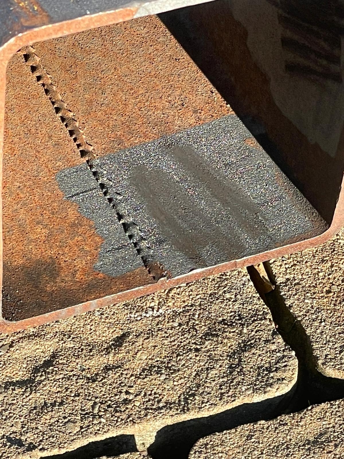 Fragment zardzewiałego, metalowego profilu z widocznym śladem cięcia i obszarem po piaskowaniu, leżącego na piasku. Detal obróbki metalu.