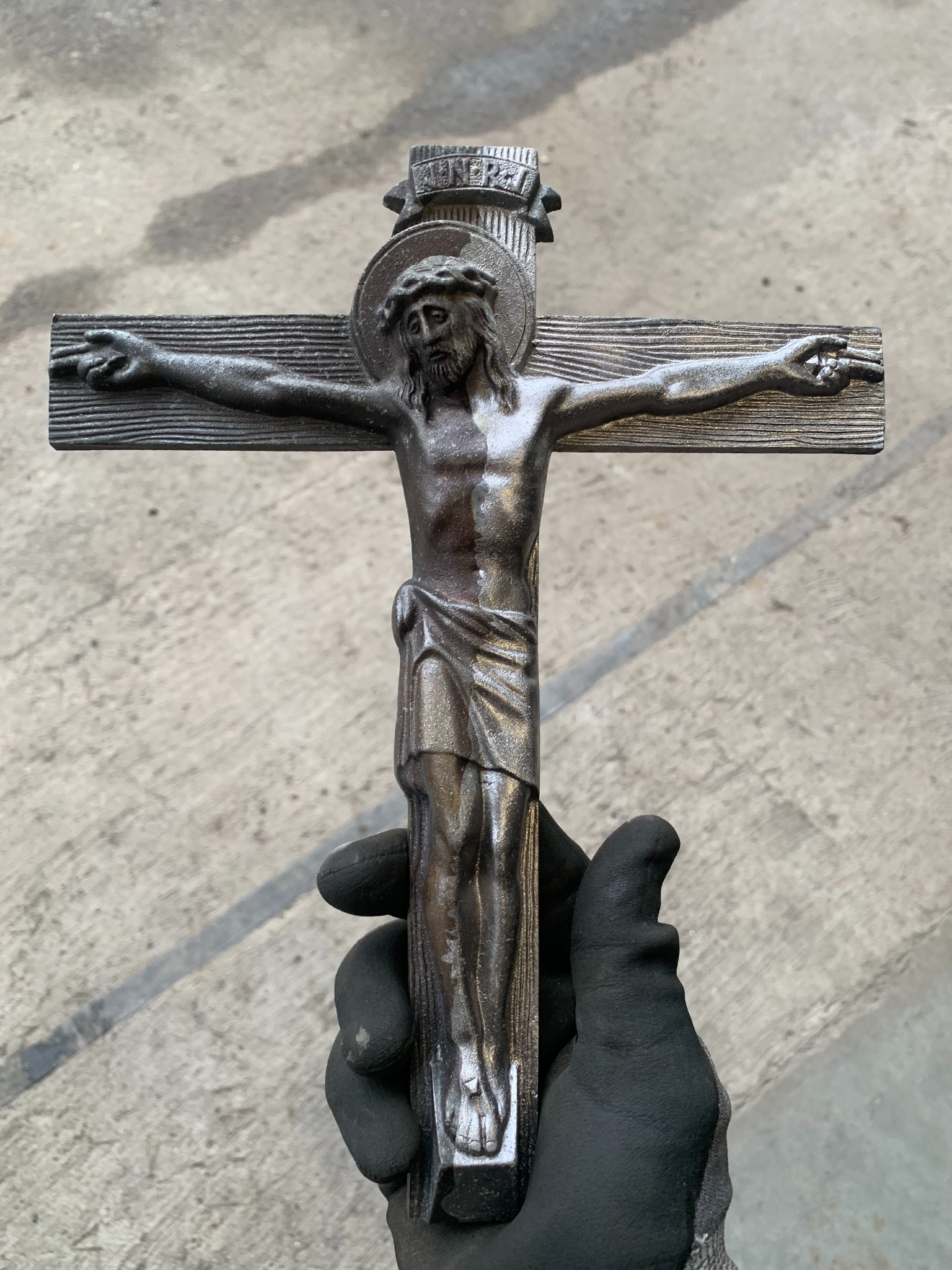 Metalowy krucyfiks trzymany w rękawicy roboczej na tle betonowej posadzki. Widoczne detale faktury metalu i zarys postaci Jezusa.