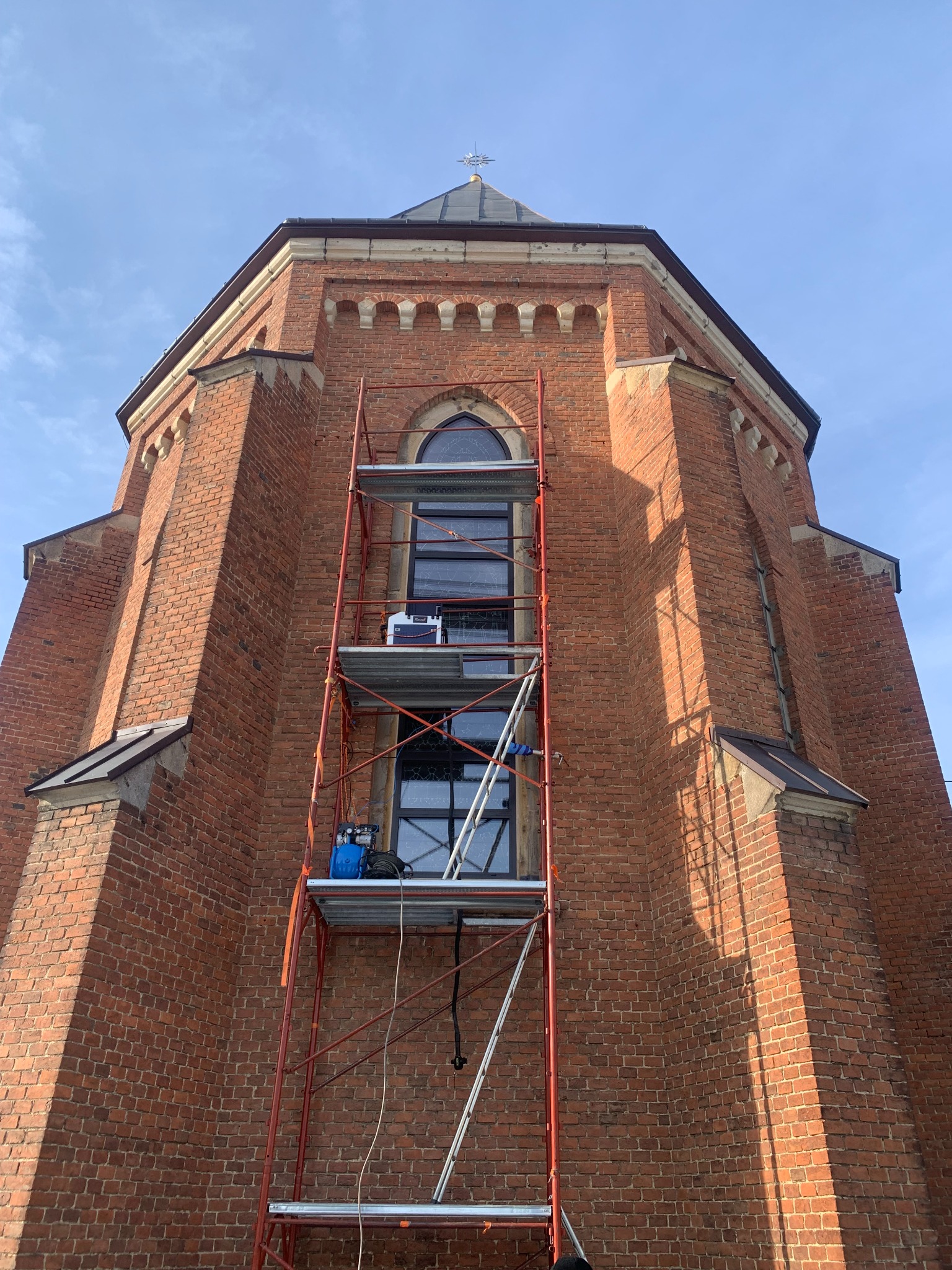 Ceglana fasada kościoła w trakcie renowacji, z rusztowaniem i sprzętem do czyszczenia elewacji. Widok z dołu, błękitne niebo w tle.