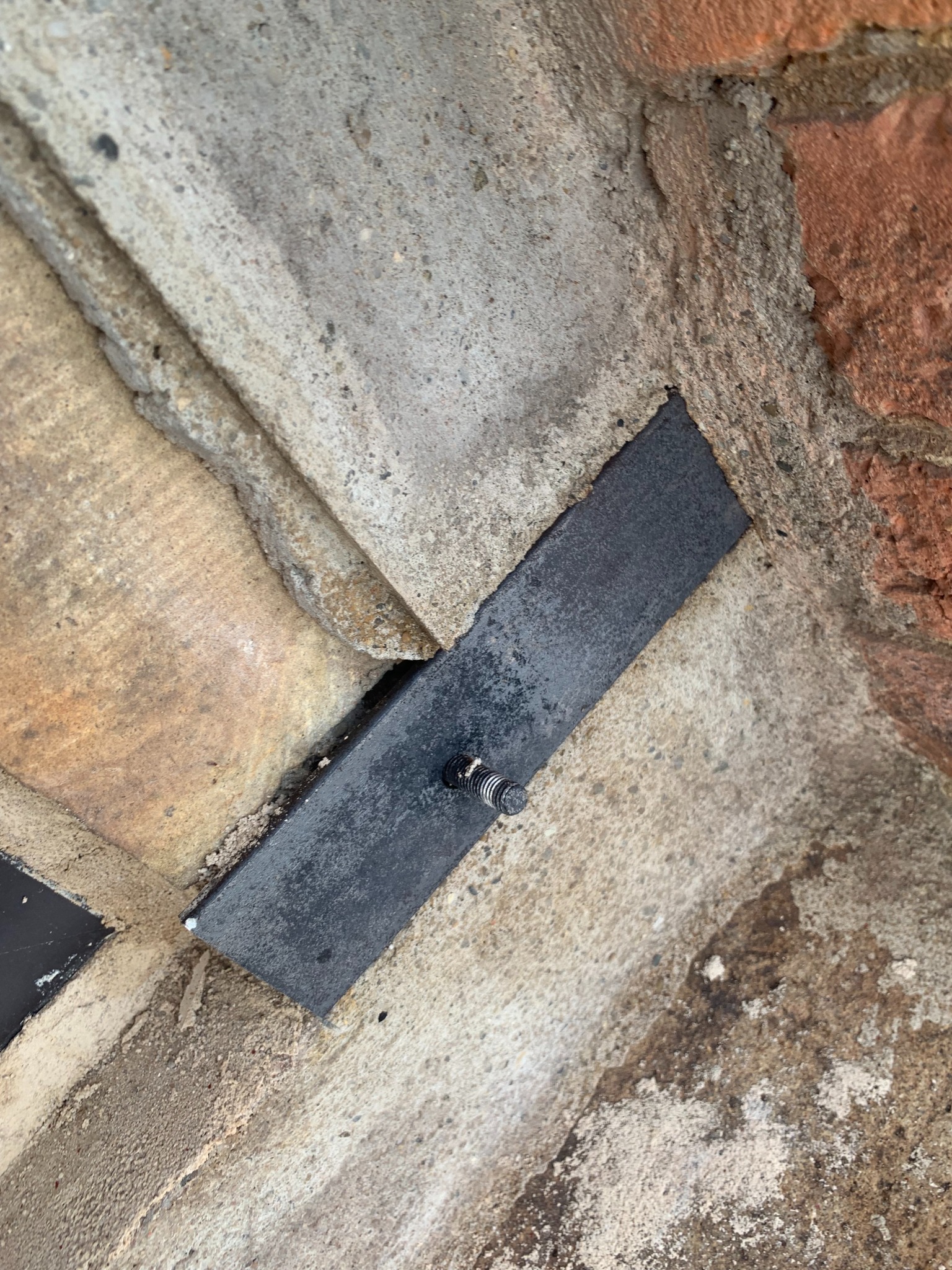 Metalowa płytka z śrubą mocująca, wkomponowana w nieregularną powierzchnię z betonu i cegieł. Detal konstrukcyjny z bliska, ukazujący połączenie materiałów.