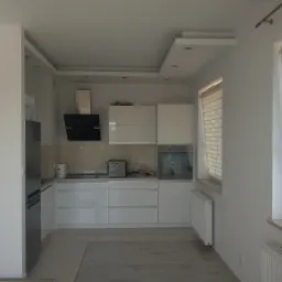 Nowoczesna, biała kuchnia z błyszczącymi frontami, czarnym okapem i oświetleniem punktowym na suficie. Widoczny fragment okna z żaluzjami i grzejnik pod oknem.