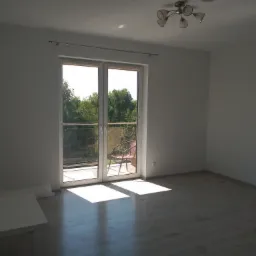 Pusty pokój z nowymi panelami podłogowymi, białymi ścianami i balkonem z widokiem na zieleń, jasne promienie słońca wpadają przez drzwi balkonowe.