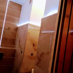 Wykończona łazienka z drewnopodobnymi płytkami, podświetleniem LED wzdłuż sufitu i marmurkowymi panelami prysznicowymi, widok zza drewnianej ościeżnicy.